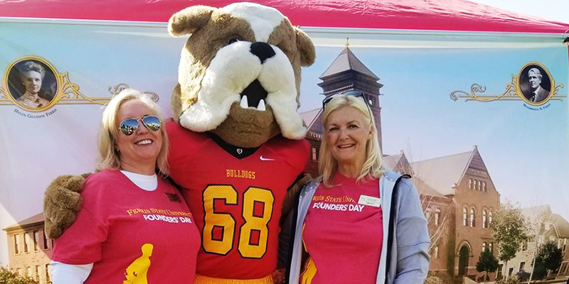 Tammy Smith and Julie Ferris pose with Ferris' mascot, Brutus, in front of an image of Ferris' Old Main building during Ferris' 2017 Founders Day.